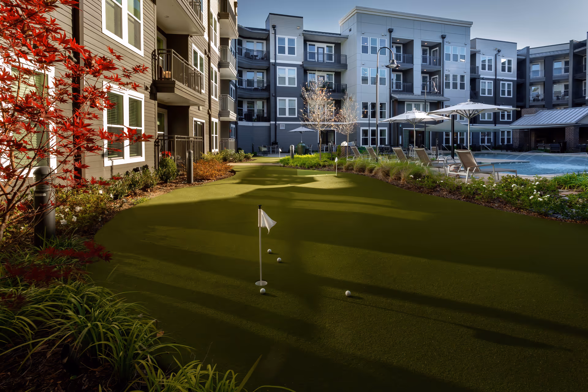Outdoor courtyard of a senior living facility featuring a putting green with golf balls and flags, surrounded by landscaped plants and trees. In the background, there is a swimming pool with lounge chairs and umbrellas, and multi-story residential buildings with balconies.