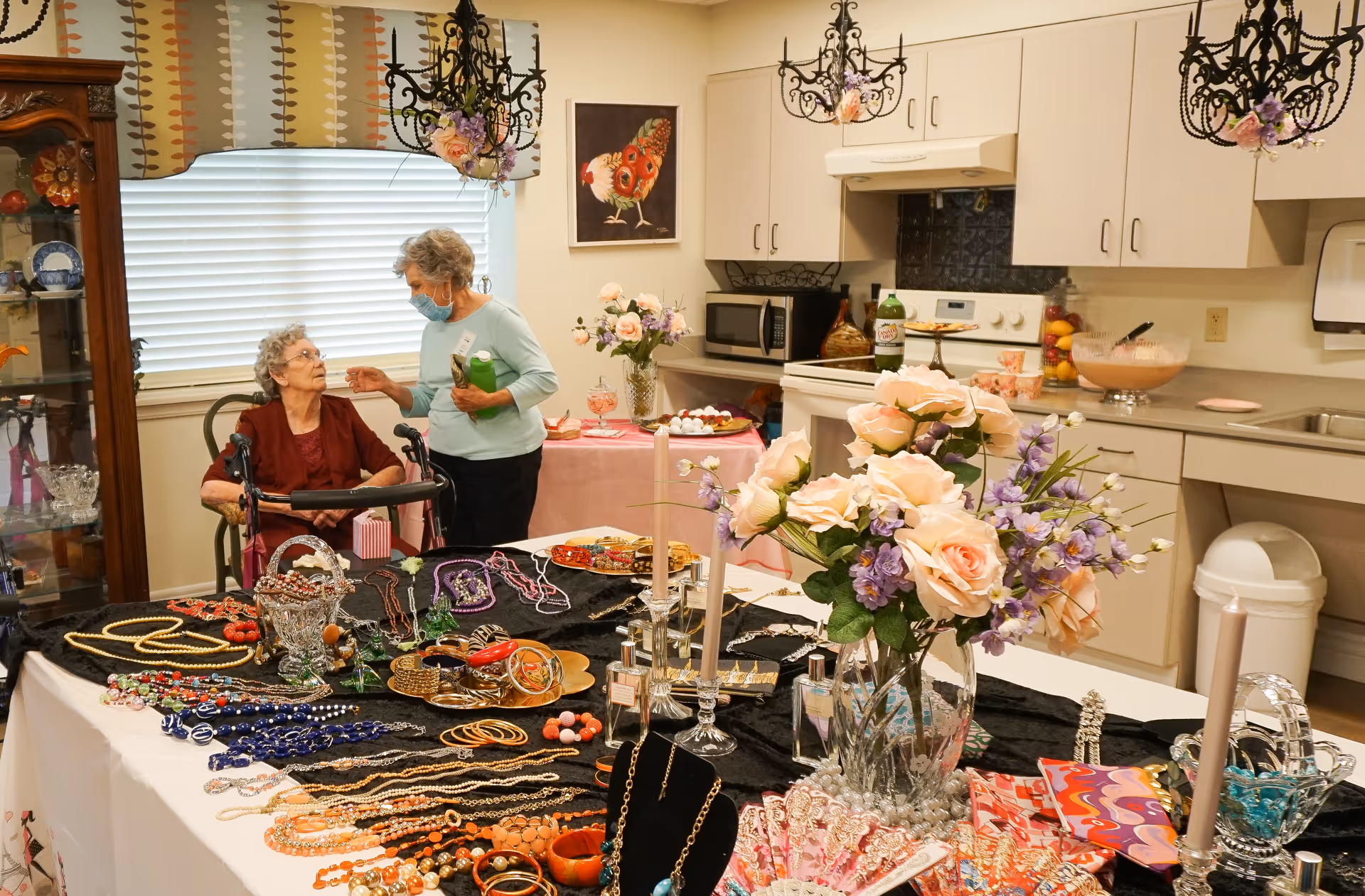 Two elderly women in a kitchen area of a senior living facility. One woman is seated with a walker, and the other is standing and talking to her while holding a green bottle. The kitchen counter and table are decorated with various jewelry pieces, flowers in vases, candles, and other decorative items. The room has beige cabinets, a microwave, stove, and a window with blinds and a patterned valance.