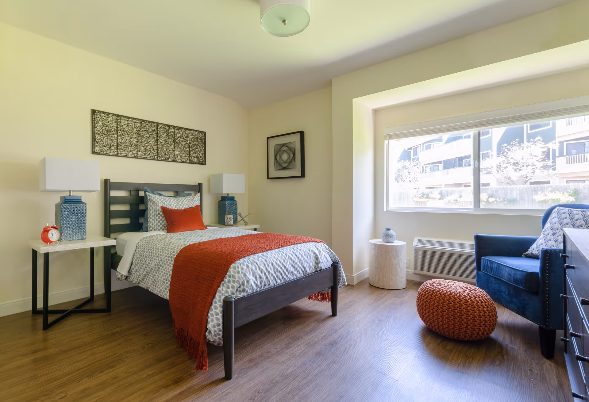 A bright bedroom with a single bed featuring patterned bedding and a red throw blanket. There are two bedside tables with blue lamps and a red alarm clock on one side. A large window lets in natural light, and a blue armchair with a patterned pillow sits near the window next to a small round table and an orange knitted pouf on the wooden floor. The walls are decorated with two framed artworks.