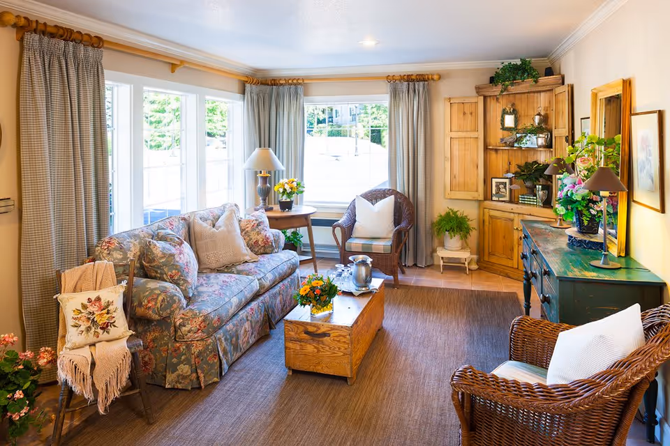 A cozy living room with a floral patterned sofa, wicker chairs with white cushions, a wooden coffee table with a flower arrangement and a silver pitcher, a side table with a lamp and flowers, large windows with curtains letting in natural light, and wooden furniture including a cabinet and a green dresser decorated with plants and framed pictures.