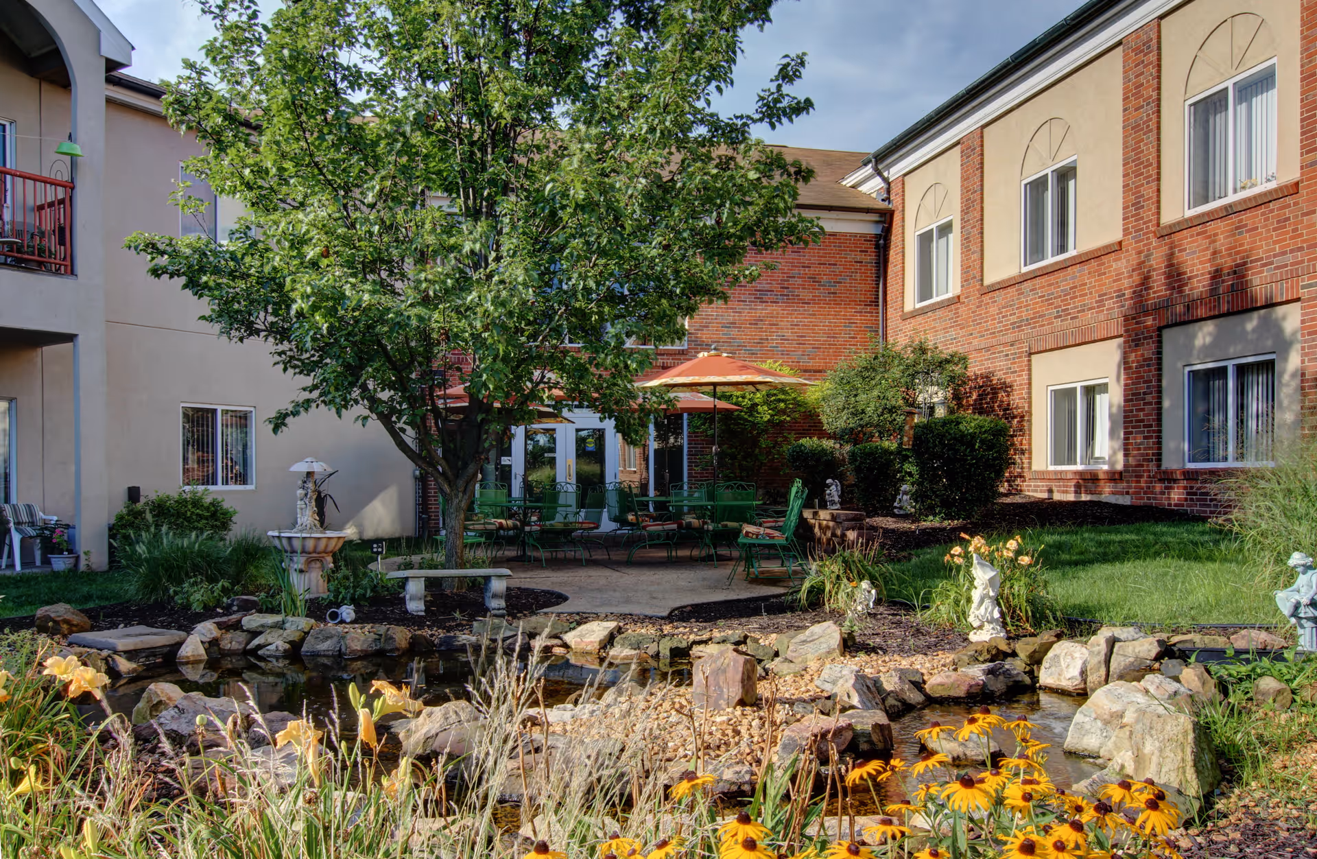 Outdoor courtyard area at Richmond Terrace featuring a small pond with rocks and yellow flowers in the foreground, a tree providing shade, green patio chairs and tables with umbrellas, and a brick building with windows in the background.