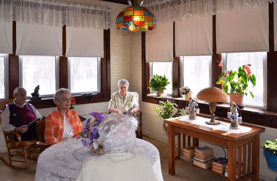A cozy sunroom with four elderly people seated around a round table covered with a lace tablecloth and a flower centerpiece. The room has multiple windows with white blinds and lace curtains, a colorful stained glass hanging lamp, and a wooden side table with potted plants, decorative figurines, a lamp, and books underneath.