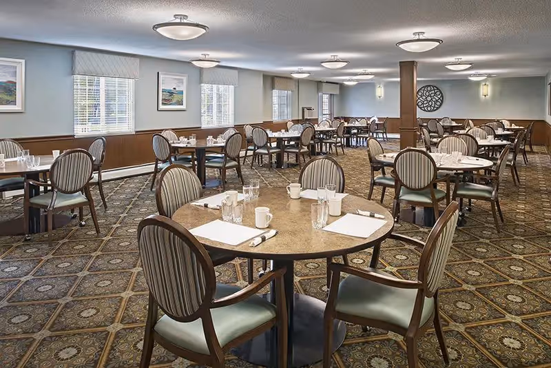 A spacious dining room with multiple round and rectangular tables set with white napkins, glasses, and coffee mugs. The room has patterned carpet flooring, light blue walls with wooden paneling, several windows with blinds, and ceiling lights providing illumination.