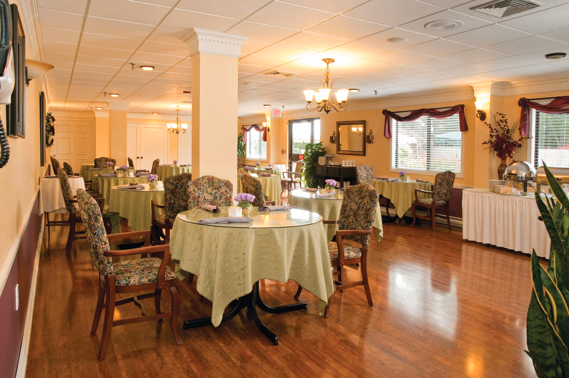 Bright, well-appointed dining room with round tables covered in light green tablecloths, floral upholstered chairs, chandeliers, and a buffet along the wall.