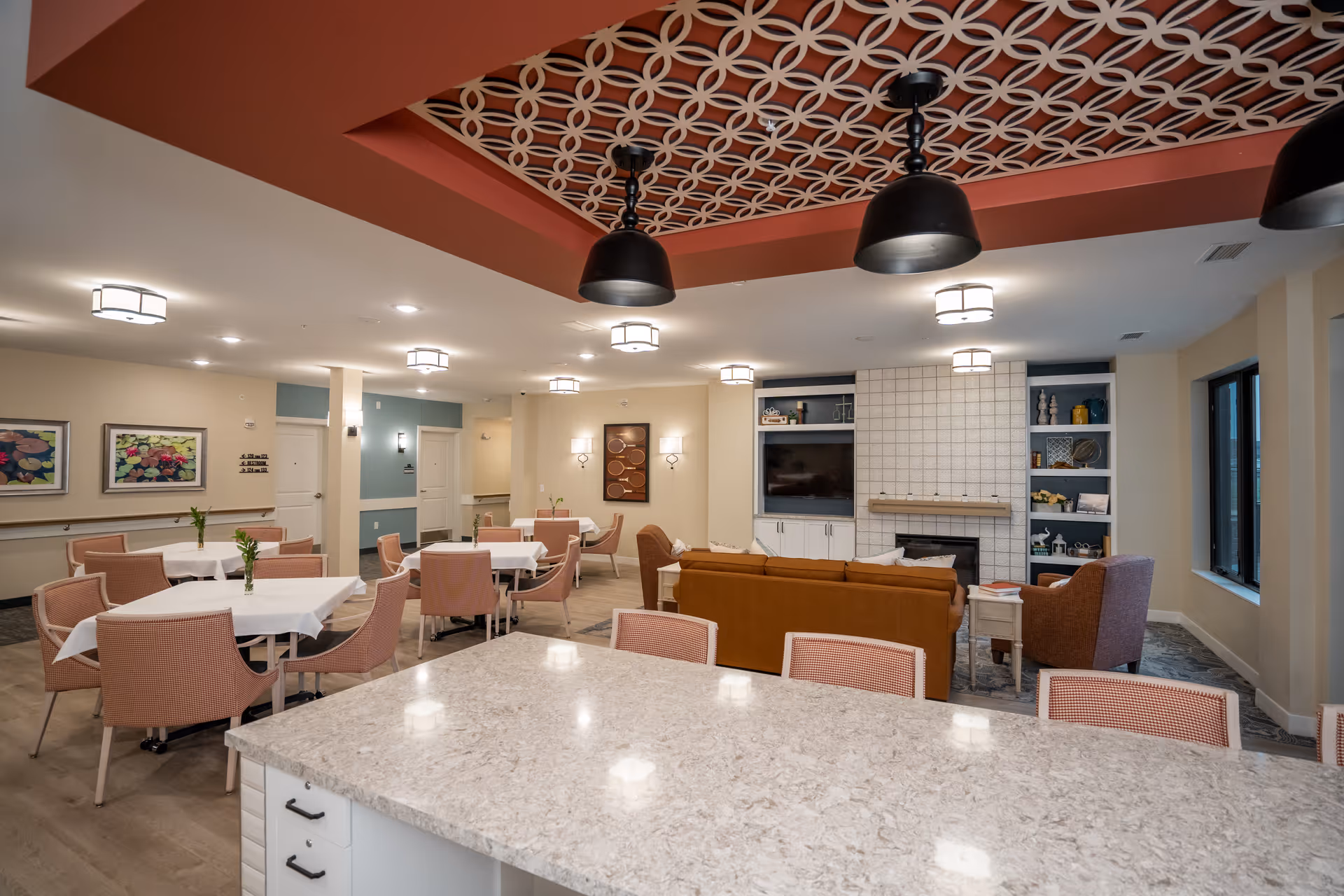 Communal dining and lounge area with multiple tables and pink chairs, a sofa facing a fireplace/TV, and decorative patterned ceiling panels.
