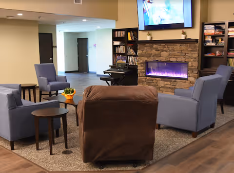 Communal living room with upholstered chairs arranged around a stone electric fireplace and wall-mounted TV.