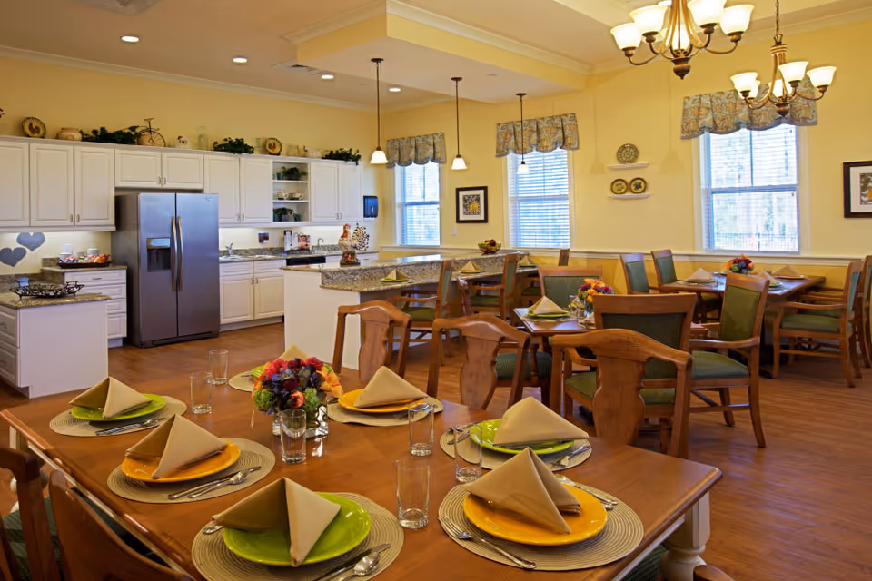 A bright dining room with wooden tables and chairs set with colorful plates, folded napkins, glasses, and floral centerpieces. The room features large windows with patterned valances, a kitchen area with white cabinets, granite countertops, and stainless steel refrigerator. Warm yellow walls and chandeliers provide a cozy atmosphere.