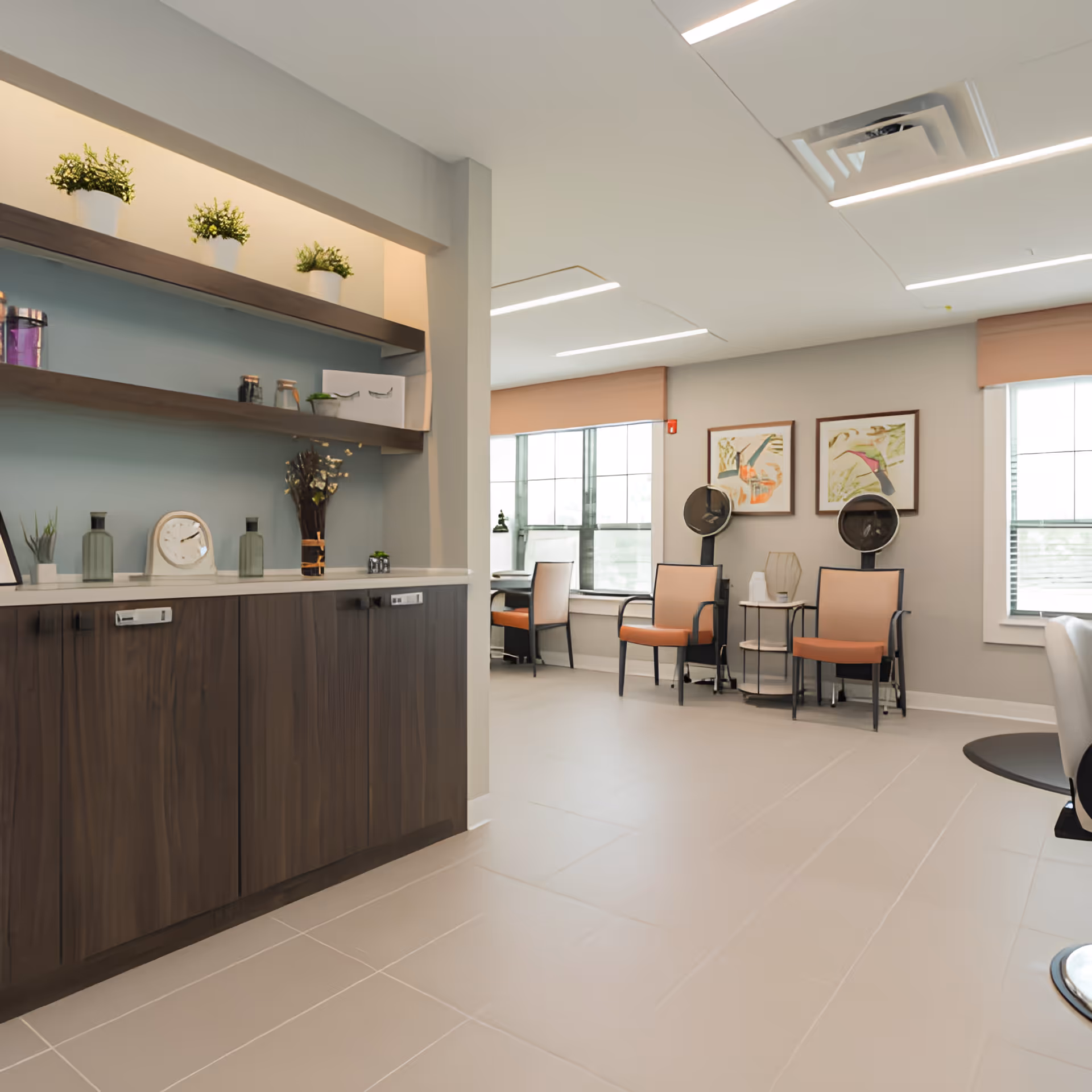A bright and modern interior room with large windows, featuring two hair drying chairs with hooded dryers, orange cushioned chairs, framed abstract artwork on the wall, and a wooden cabinet with shelves holding small plants and decorative items.