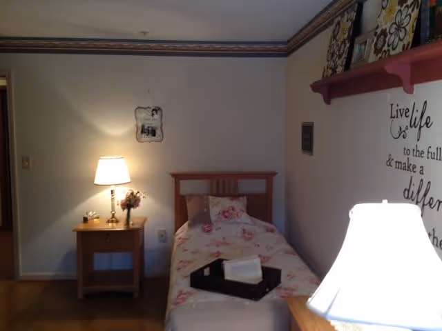 A small bedroom with a single bed covered in floral bedding. There is a wooden nightstand next to the bed with a lit table lamp and a small flower arrangement. On the wall above the bed, there is a decorative shelf with framed pictures and a wall decal that reads 'Live life to the full & make a difference.' The room has wooden flooring and a wallpaper border near the ceiling.