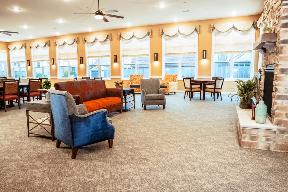 A spacious and well-lit common area with multiple seating options including armchairs, a sofa, and dining tables with chairs. Large windows line the back wall, allowing natural light to fill the room. A stone fireplace with decorative vases and plants is visible on the right side. The room has a carpeted floor and ceiling fans.
