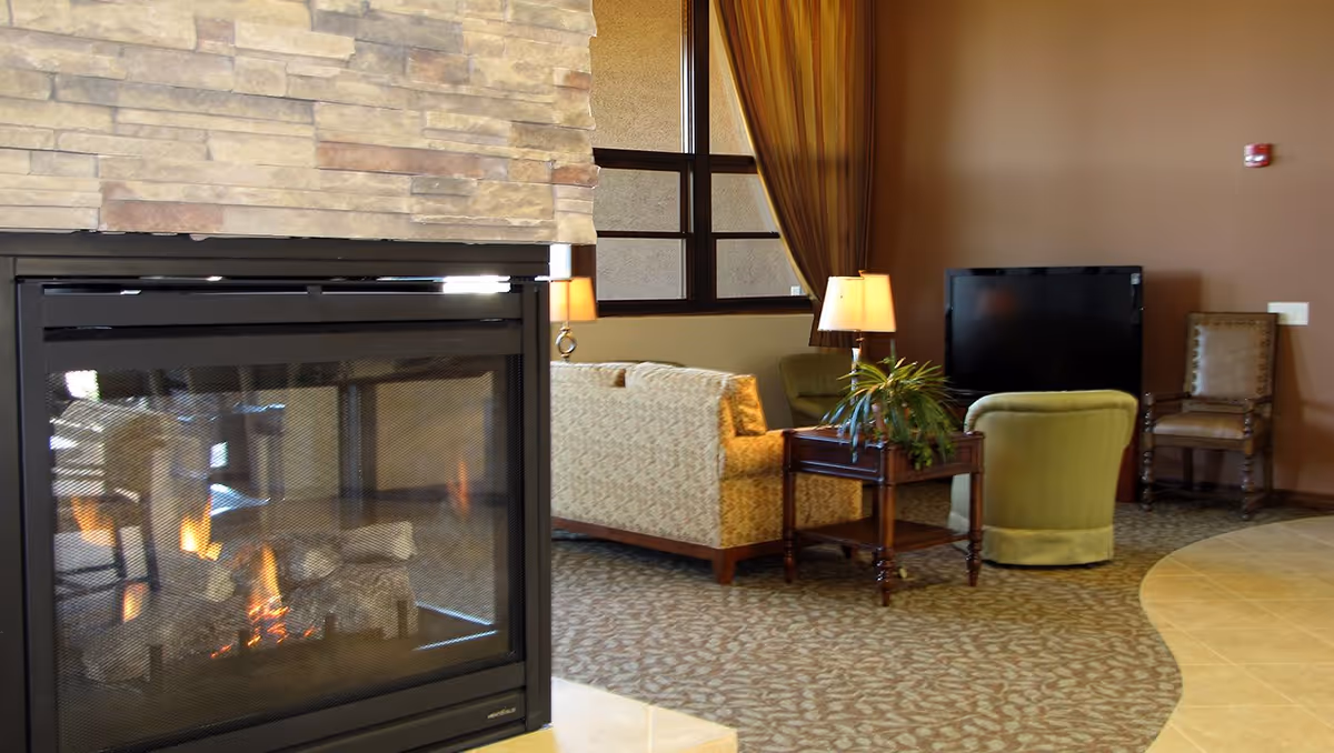 A cozy communal living room with a lit fireplace, upholstered sofas and chairs, a side table with a lamp and plant, and a TV.