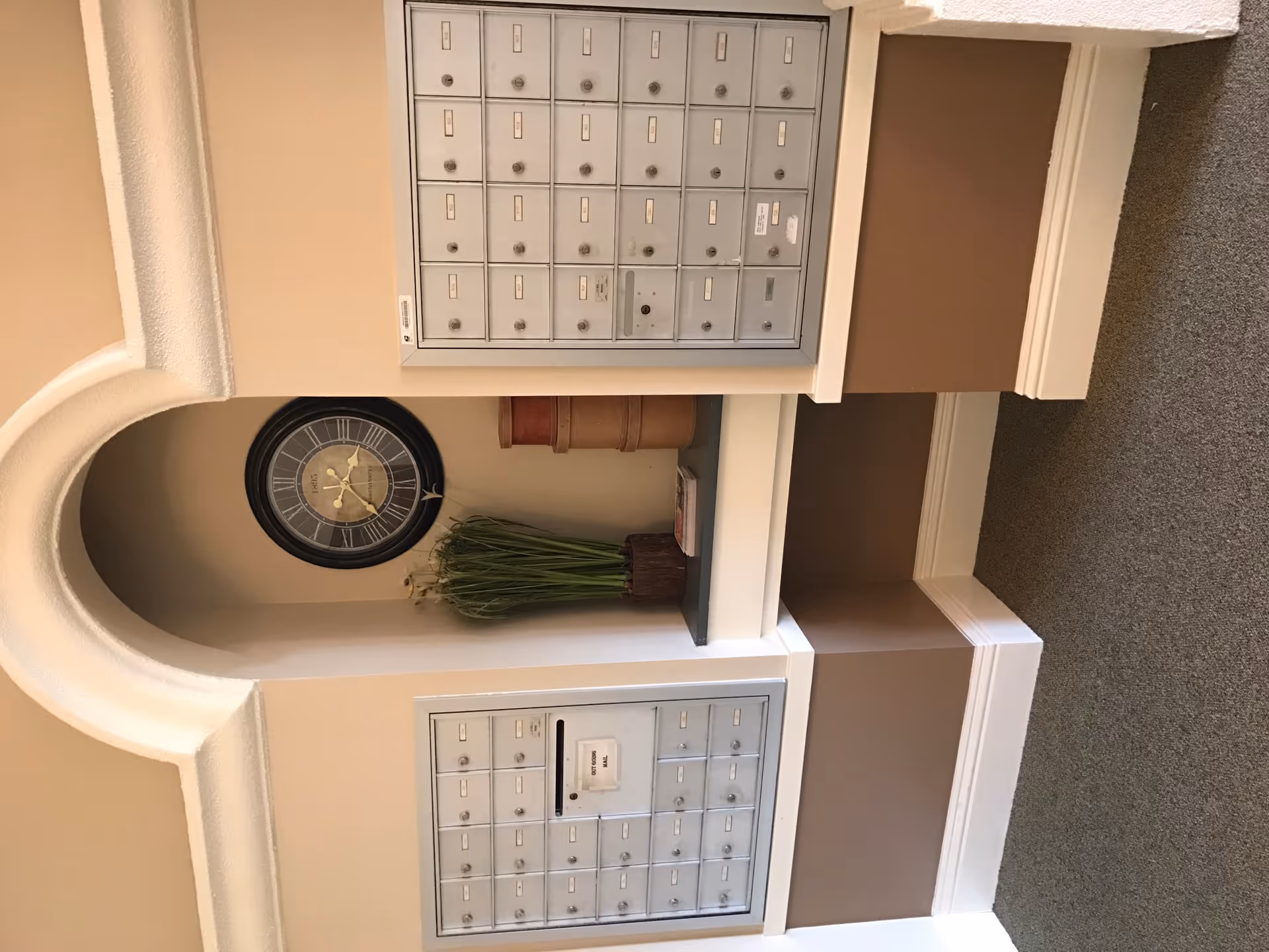 Interior view of a mail area with two sets of silver mailboxes mounted on a beige wall. Between the mailboxes is a decorative alcove containing a large round clock, a tall vase with green plants, and a small stack of books. The floor is carpeted in gray.