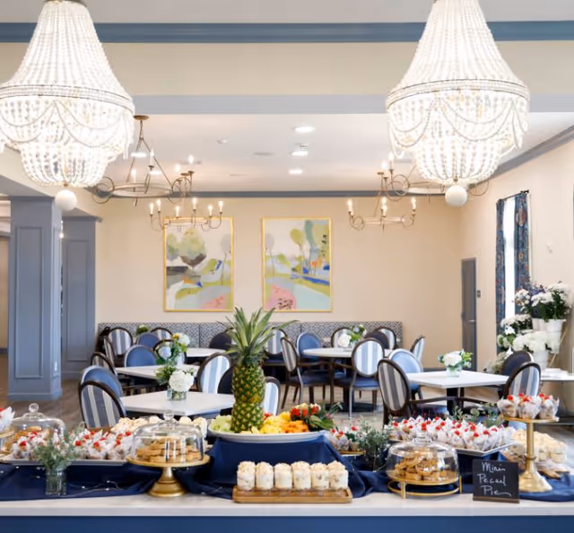 A bright and elegant dining room with round tables and chairs arranged neatly. The tables have small floral centerpieces. In the foreground, a buffet table is set with various desserts, including mini pecan pies, cookies, and cupcakes, with a pineapple centerpiece. The room features large chandeliers, modern light fixtures, abstract paintings on the wall, and floral arrangements near the windows.