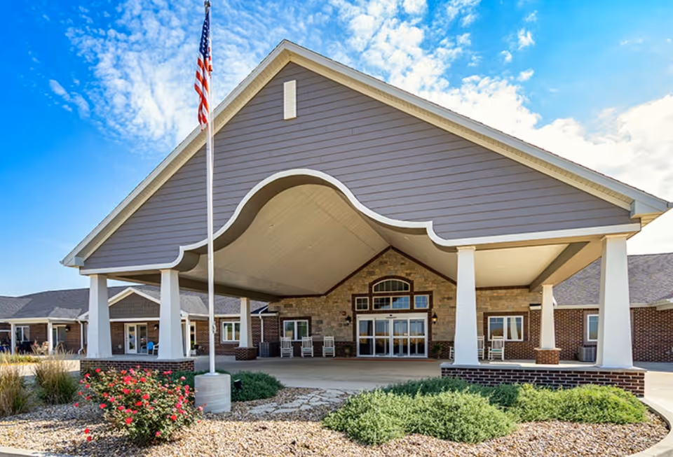 Front exterior view of a senior living facility with a large covered entrance supported by white columns, an American flag on a flagpole, landscaped bushes and flowers, and a clear blue sky with some clouds.