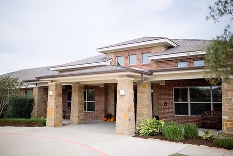 Exterior view of a single-story brick building with stone pillars supporting a covered entrance. There are windows, plants, and a bench near the entrance under a partly cloudy sky.