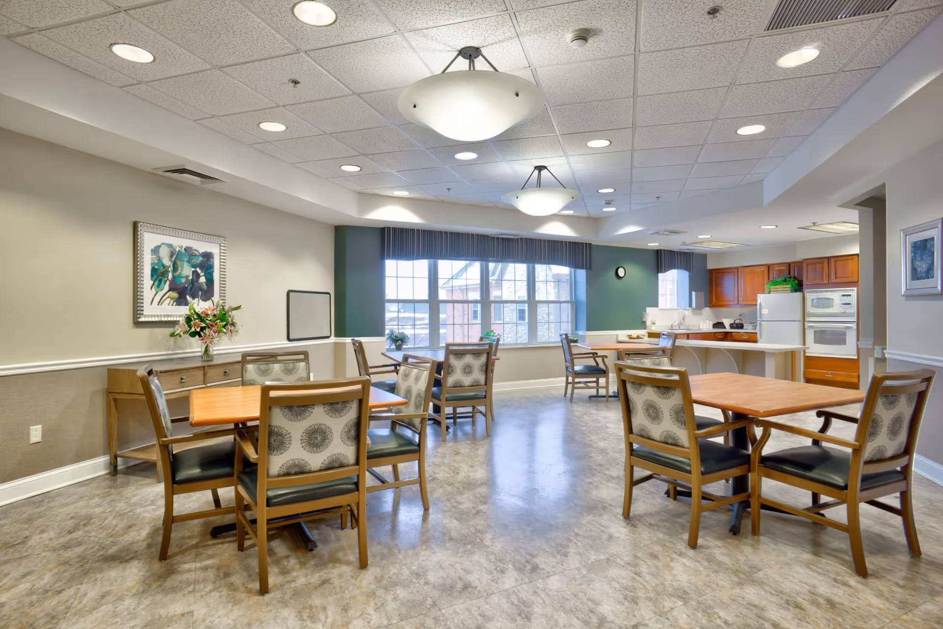 A bright dining area in a senior living facility with several wooden tables and cushioned chairs arranged neatly. The room features large windows with valances allowing natural light to fill the space. There is a kitchen area in the background with wooden cabinets, a refrigerator, microwave, and oven. The walls are painted in neutral tones with some artwork and a flower arrangement on a side table.