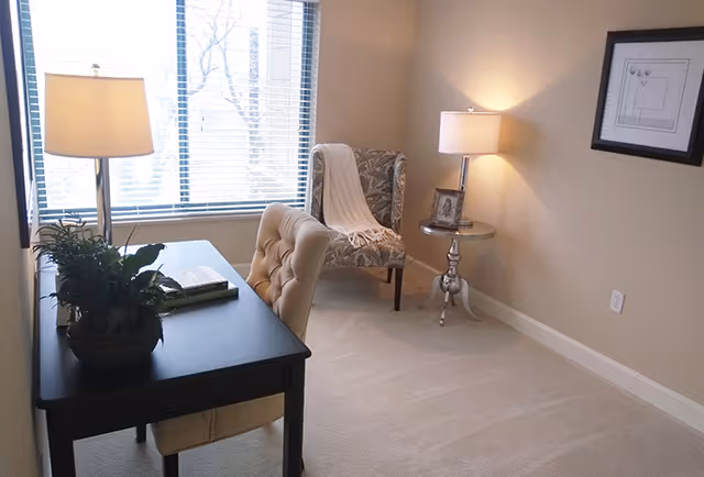 A small, cozy room with a black desk and a beige tufted chair. On the desk, there is a potted plant, a table lamp, and a book. Near the window with blinds, there is an upholstered armchair with a white throw draped over it. A round side table with a lamp and a framed photo is positioned next to the armchair. The walls are beige, and a framed picture hangs on the right wall.