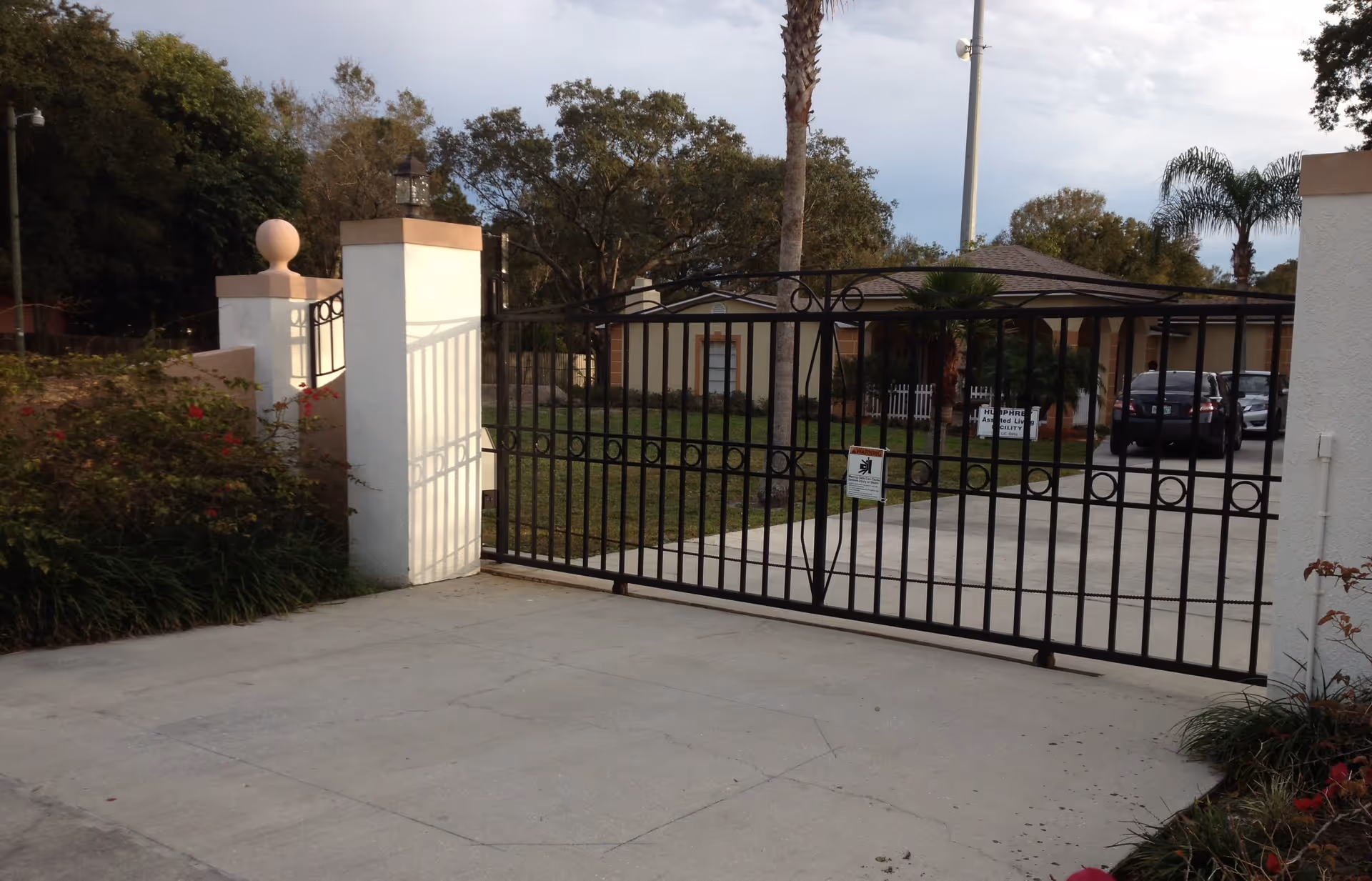 Black metal gate at the entrance of a facility with a driveway leading to a building in the background. The building is surrounded by grass, palm trees, and other greenery. There are two cars parked near the building, and a sign indicating the facility name is visible behind the gate.