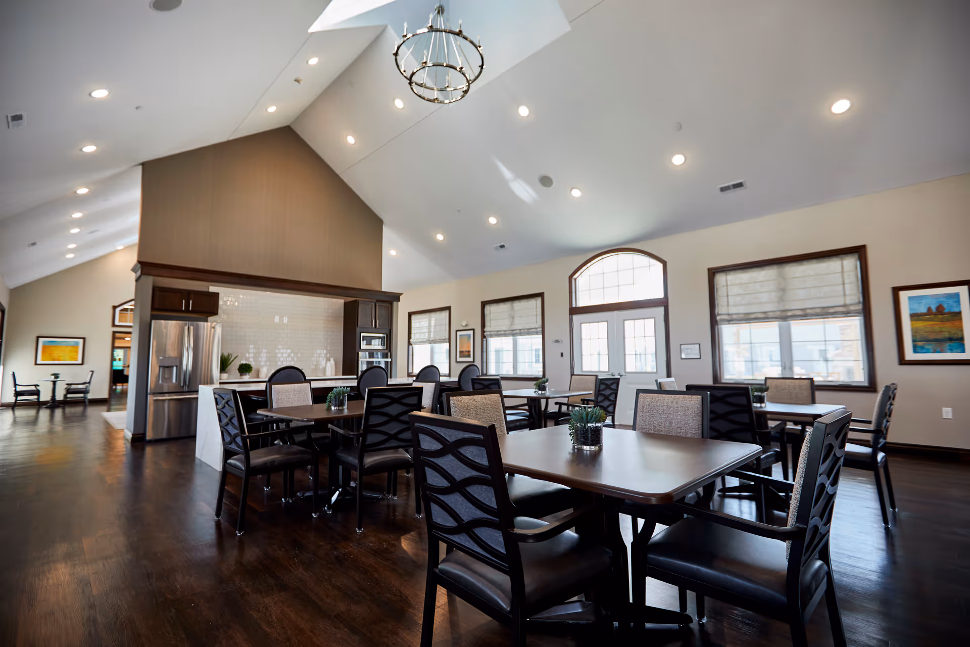 A spacious dining area in a senior living facility with multiple tables and chairs arranged neatly. The room features high vaulted ceilings with recessed lighting and a chandelier. Large windows with blinds allow natural light to fill the space. The area includes a kitchen section with stainless steel appliances and dark wood cabinetry. The walls are decorated with framed artwork.