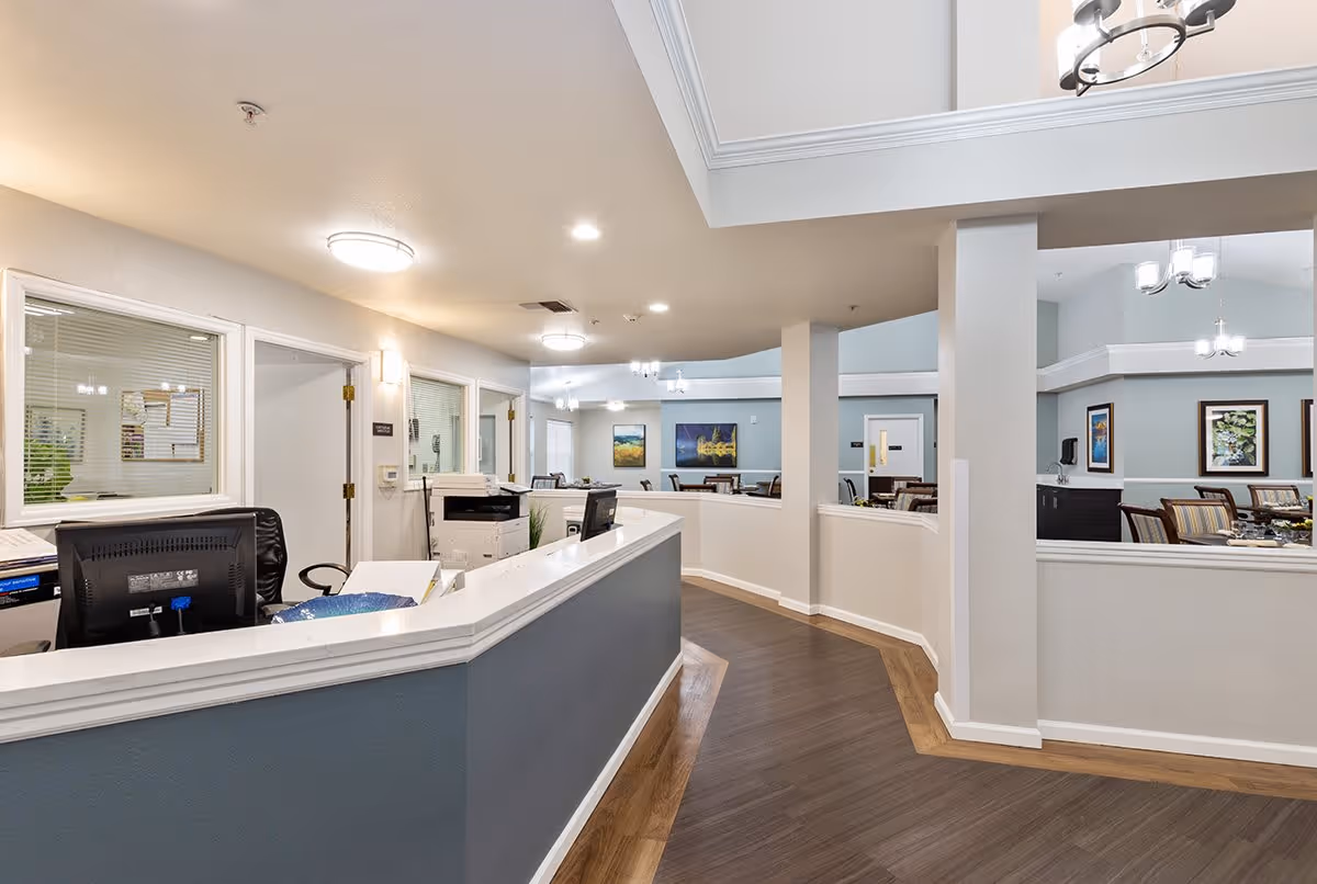 Interior view of a senior living facility reception area with a curved desk, computer monitors, and office equipment. Beyond the reception area, there is a dining or common area with tables and chairs, light blue walls, framed artwork, and ceiling light fixtures.
