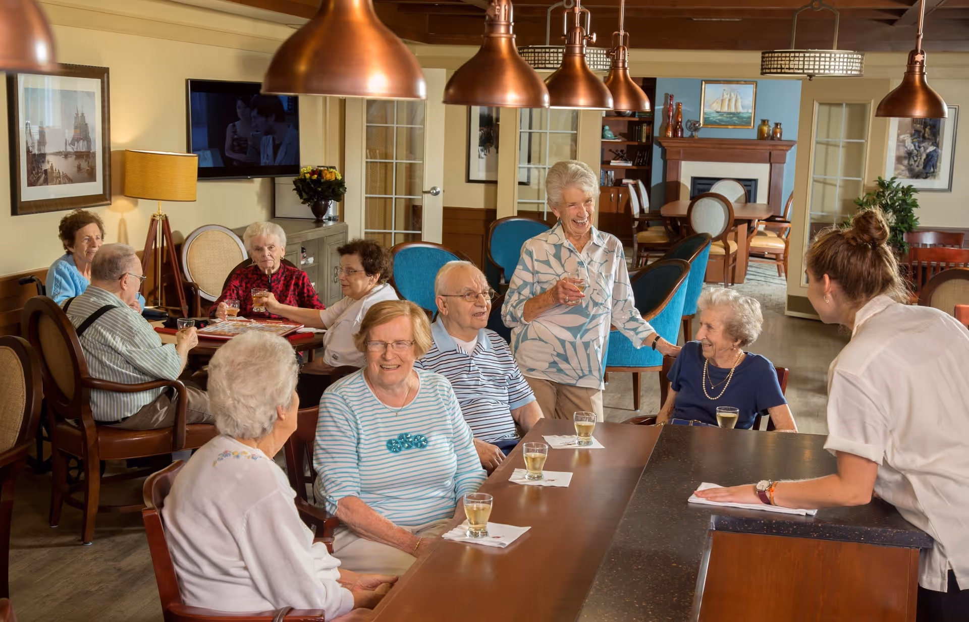 A group of elderly people socializing and enjoying drinks together in a cozy common area with wooden furniture, blue chairs, and warm lighting. A staff member is engaging with the group at a counter, and there are paintings and a TV on the walls in the background.