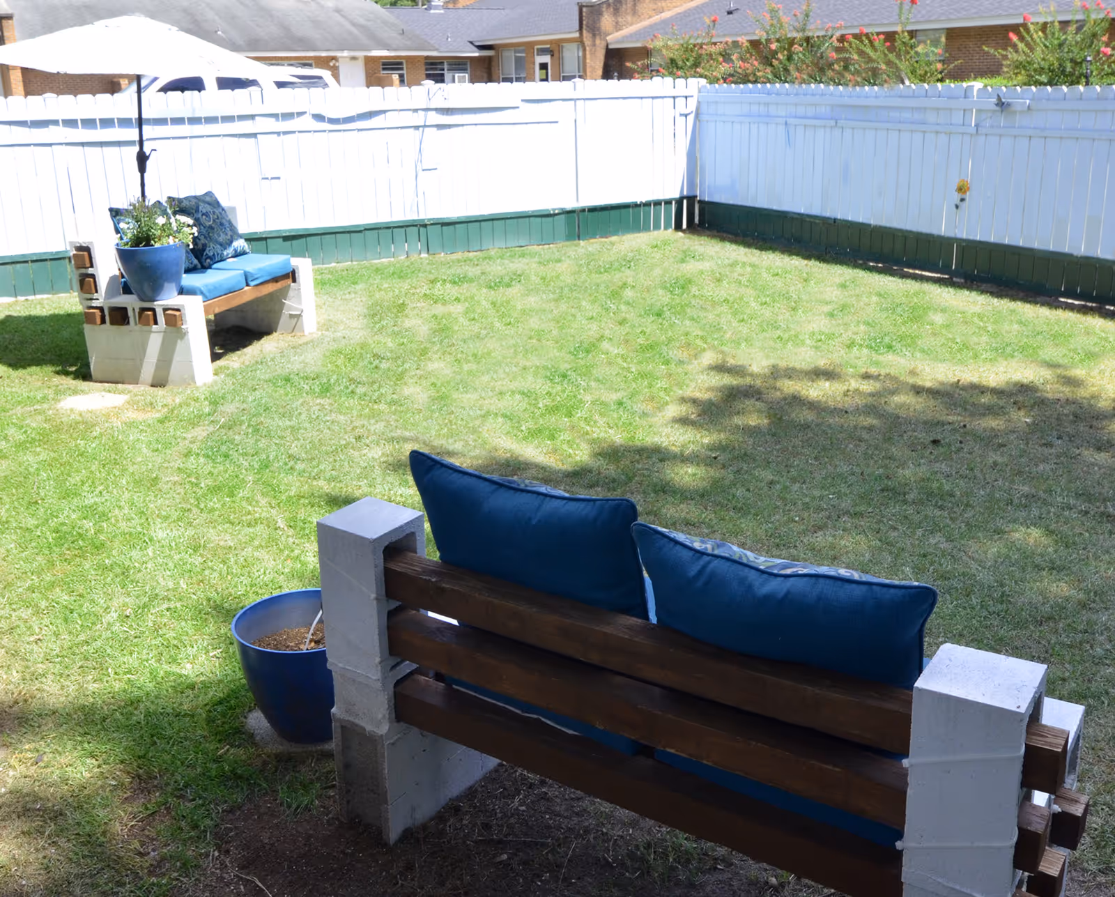 A small outdoor grassy yard enclosed by a white wooden fence with green trim. There are two benches made of wood and concrete blocks, each with blue cushions and pillows. One bench has a blue flower pot with plants on it. Some buildings and flowering bushes are visible beyond the fence.