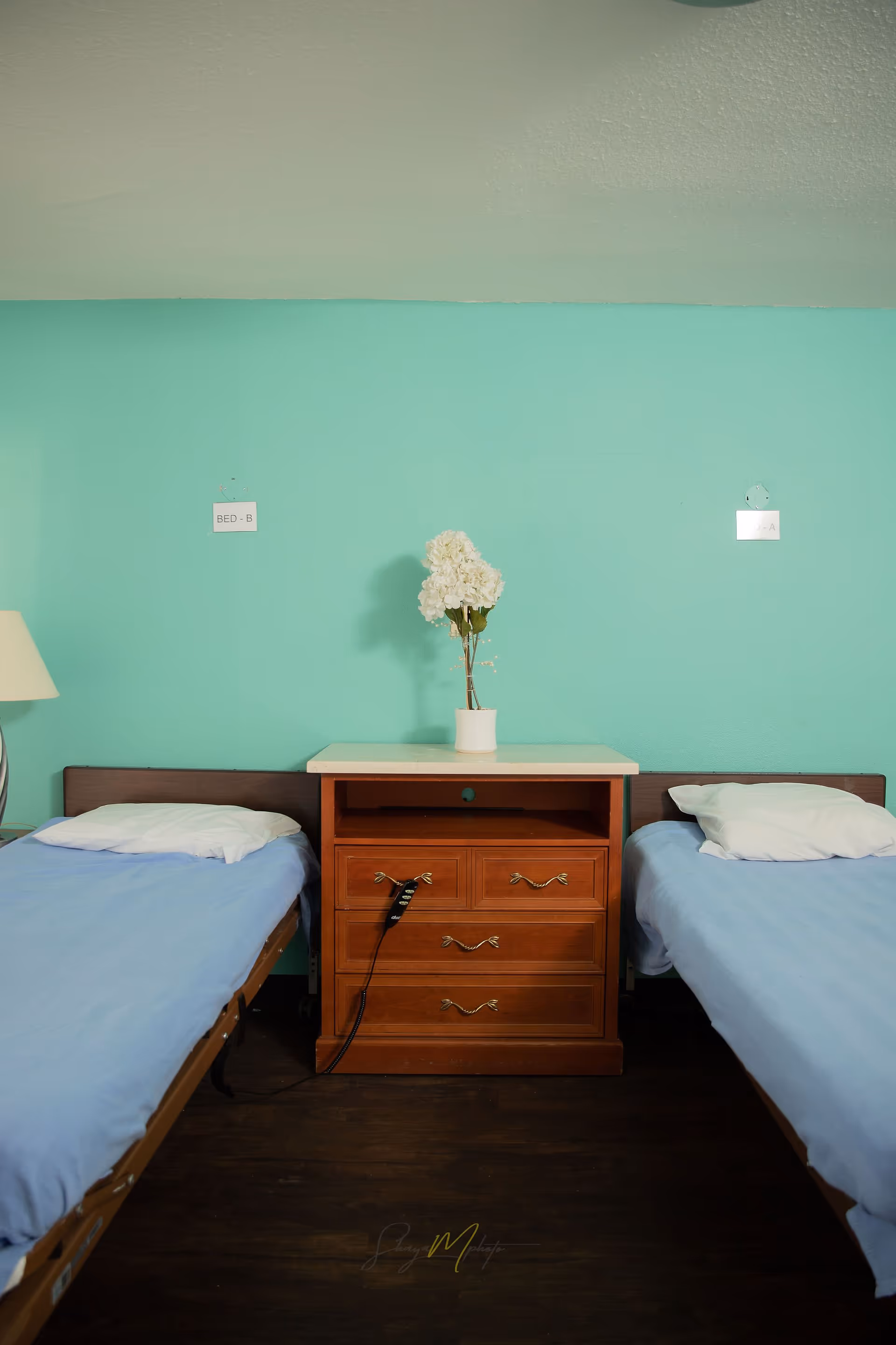 Two single beds with blue sheets and white pillows separated by a wooden nightstand with drawers. A white vase with white flowers is placed on top of the nightstand. The wall behind is painted turquoise with small signs labeled 'BED - B' and 'BED - A' above each bed.
