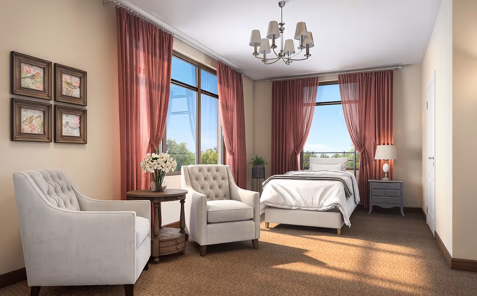 A bright and cozy bedroom with a single bed, two upholstered armchairs, a round wooden side table with a vase of flowers, large windows with red curtains letting in natural light, a bedside table with a lamp, and framed artwork on the wall.