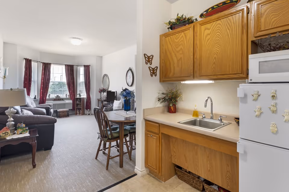 Interior view of a senior living facility apartment showing a small kitchen area with wooden cabinets, a sink, a white refrigerator with decorative magnets, and a microwave. Adjacent to the kitchen is a dining area with a round wooden table and chairs. Beyond the dining area is a living room with a sofa, armchair, side table with a lamp, and large windows with red curtains letting in natural light.