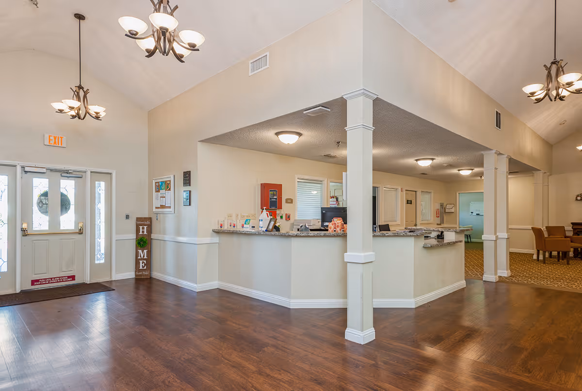 Interior view of a senior living facility reception area with a front desk, wooden flooring, beige walls, and ceiling lights. There are double glass doors with an exit sign above them, a decorative sign that says 'HOME', and a seating area with chairs visible in the background.