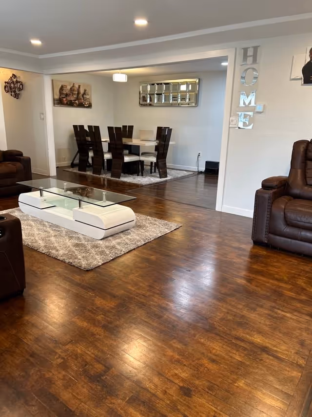 Interior view of a senior living facility showing a living room with brown leather recliners and a modern white coffee table on a gray rug. In the background, there is a dining area with a white dining table and dark wooden chairs on a rug, wall art, and a decorative mirror. The word 'HOME' is displayed vertically on the wall near the dining area.