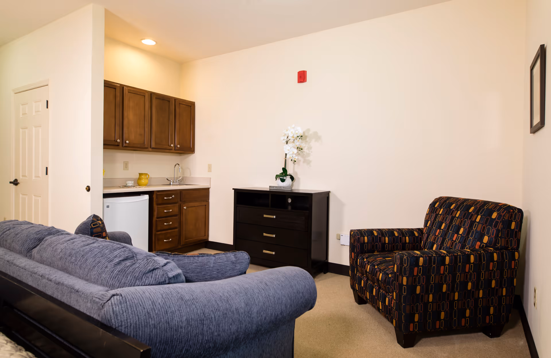 Small living area with a blue sofa, patterned armchair, kitchenette with wooden cabinets and mini-fridge, and a dark dresser topped with a white orchid.