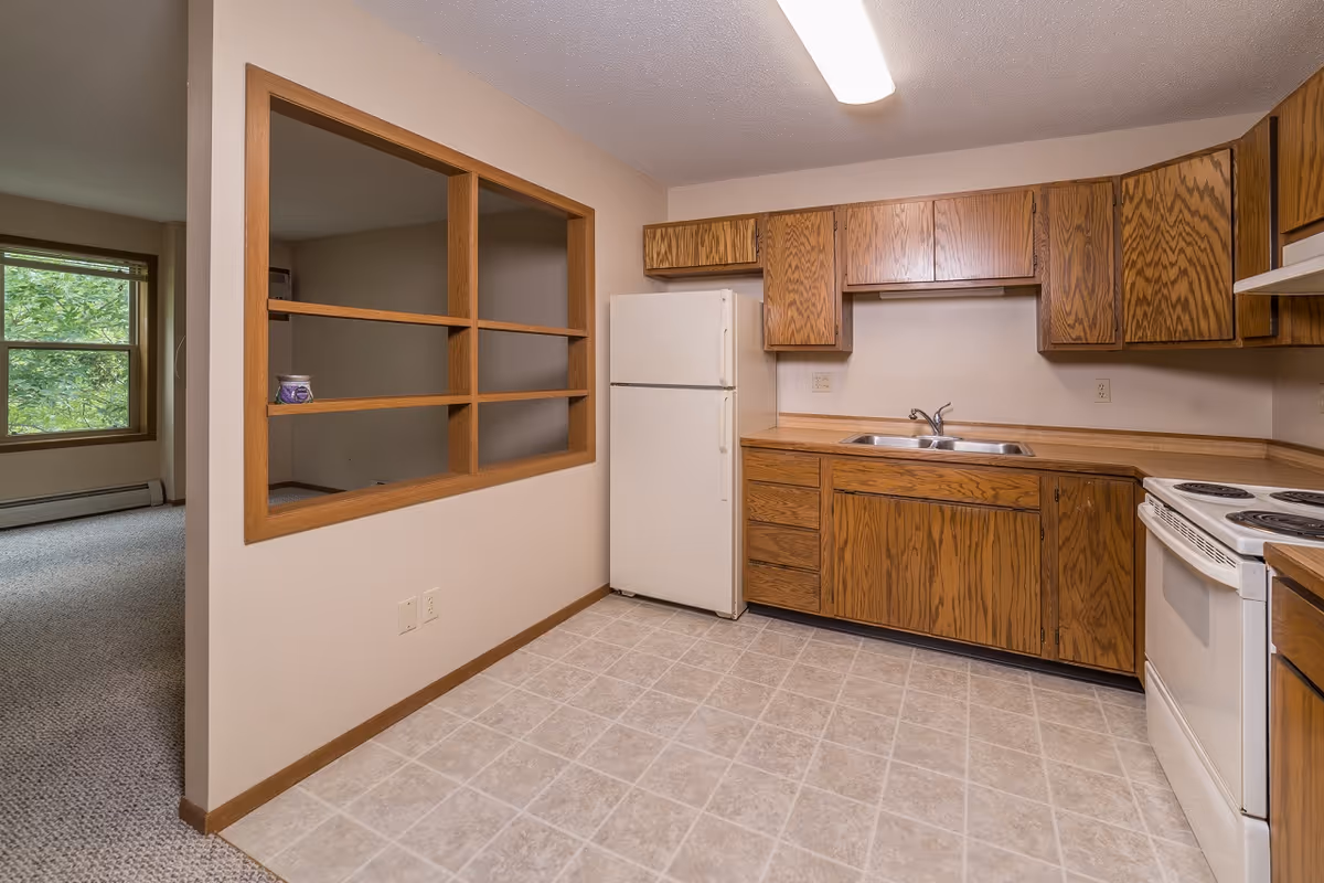 Small kitchen with wooden cabinets, a white refrigerator and stove, a double sink, tiled floor, and a pass-through window to an adjacent room.