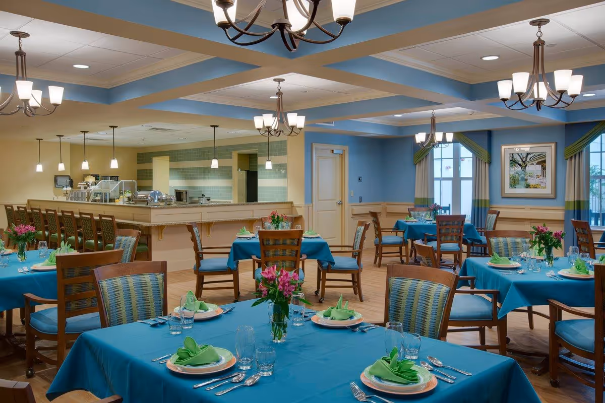A brightly lit dining room with multiple tables covered in blue tablecloths, each set with plates, silverware, glasses, and green folded napkins. There are wooden chairs with blue and green patterned cushions around the tables. The room features blue walls with beige wainscoting, several chandeliers hanging from the ceiling, and a serving counter with pendant lights in the background. A framed painting and windows with curtains are visible on the far wall.