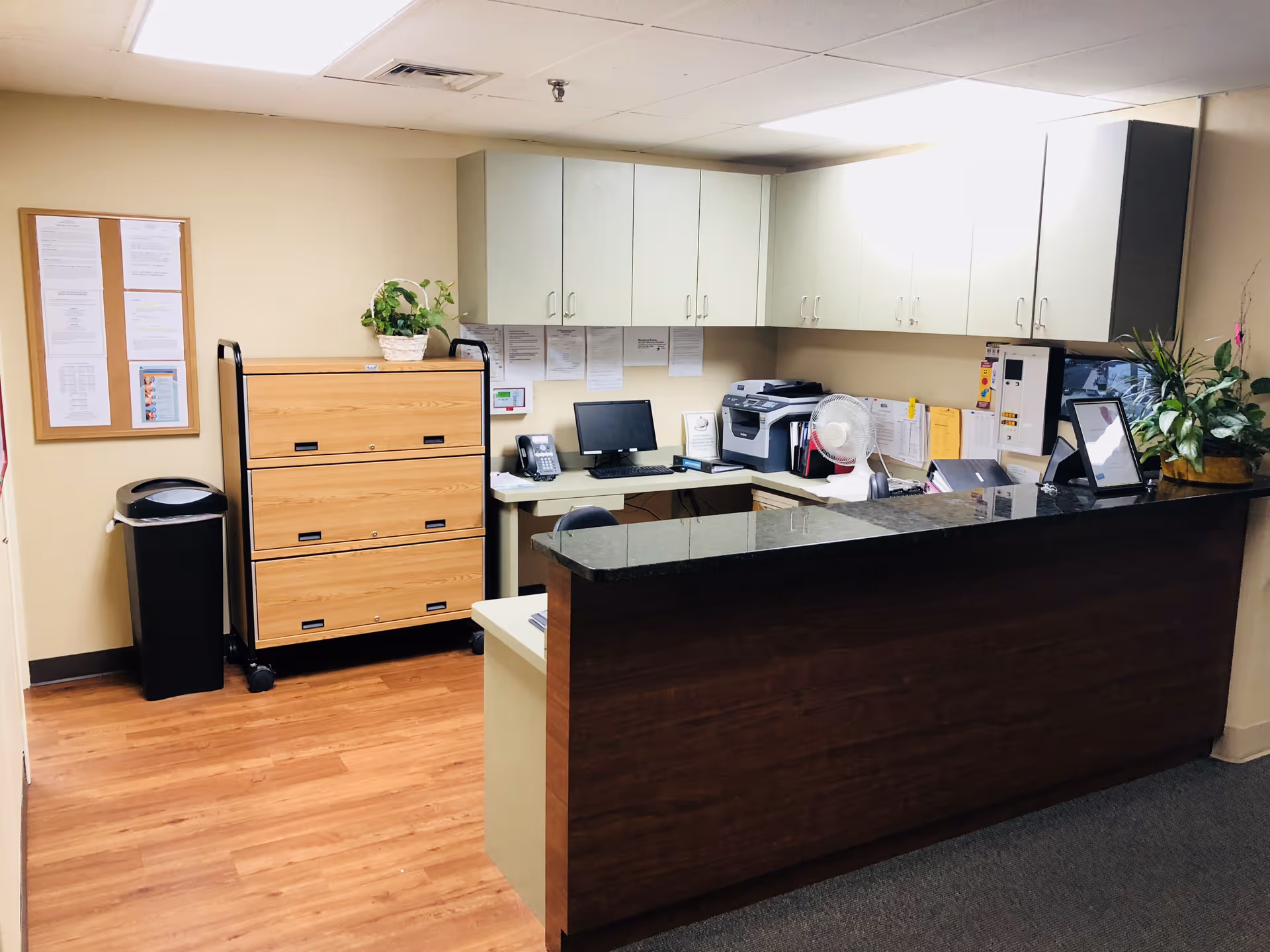 Reception desk area in a facility with a wooden counter, computer, telephone, printer, filing cabinets, bulletin board with notices, and potted plants. The floor is a mix of wood and carpet, and the walls are beige.