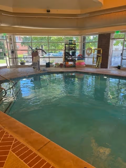 Indoor swimming pool area with clear water, surrounded by a tiled floor. Large windows in the background show greenery and parked cars outside. Pool equipment and flotation devices are visible near the windows.