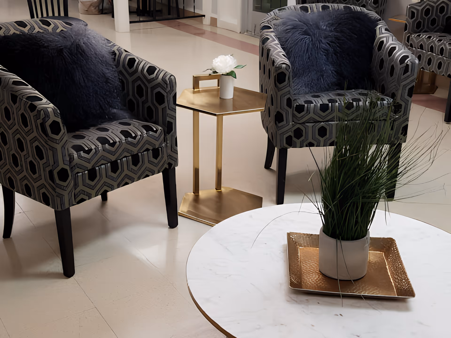 Two patterned armchairs with dark blue fluffy pillows are positioned around a small gold side table with a white flower in a vase. In the foreground, there is a round white marble table with a gold tray holding a potted green plant. The setting appears to be a clean, modern indoor common area.