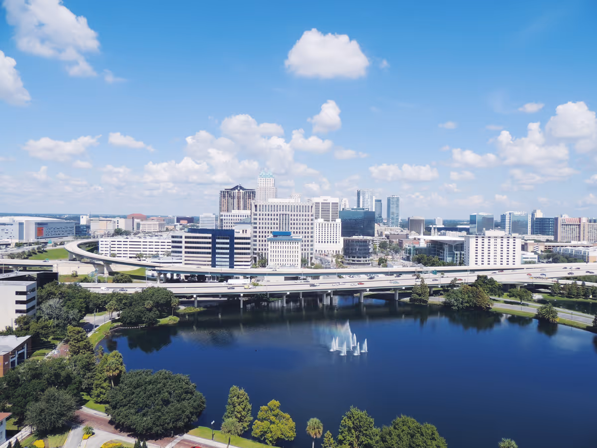 Aerial view of a downtown skyline with highways, a lake with fountains, and a bright blue sky with clouds.