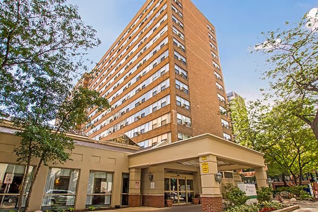 Exterior view of a multi-story brick senior living facility building with a covered entrance and surrounding trees under a clear blue sky.