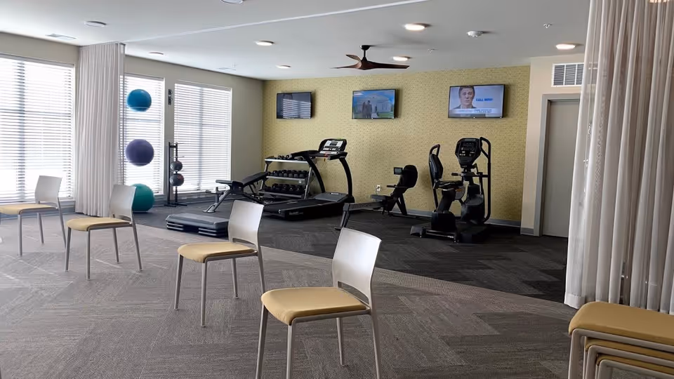 A fitness room with exercise equipment including a treadmill, stationary bikes, and dumbbells. There are three wall-mounted TVs above the equipment. In the foreground, there are several chairs arranged in a spaced-out manner, and exercise balls are stacked near the windows with blinds. The room has light-colored walls and carpeted flooring.