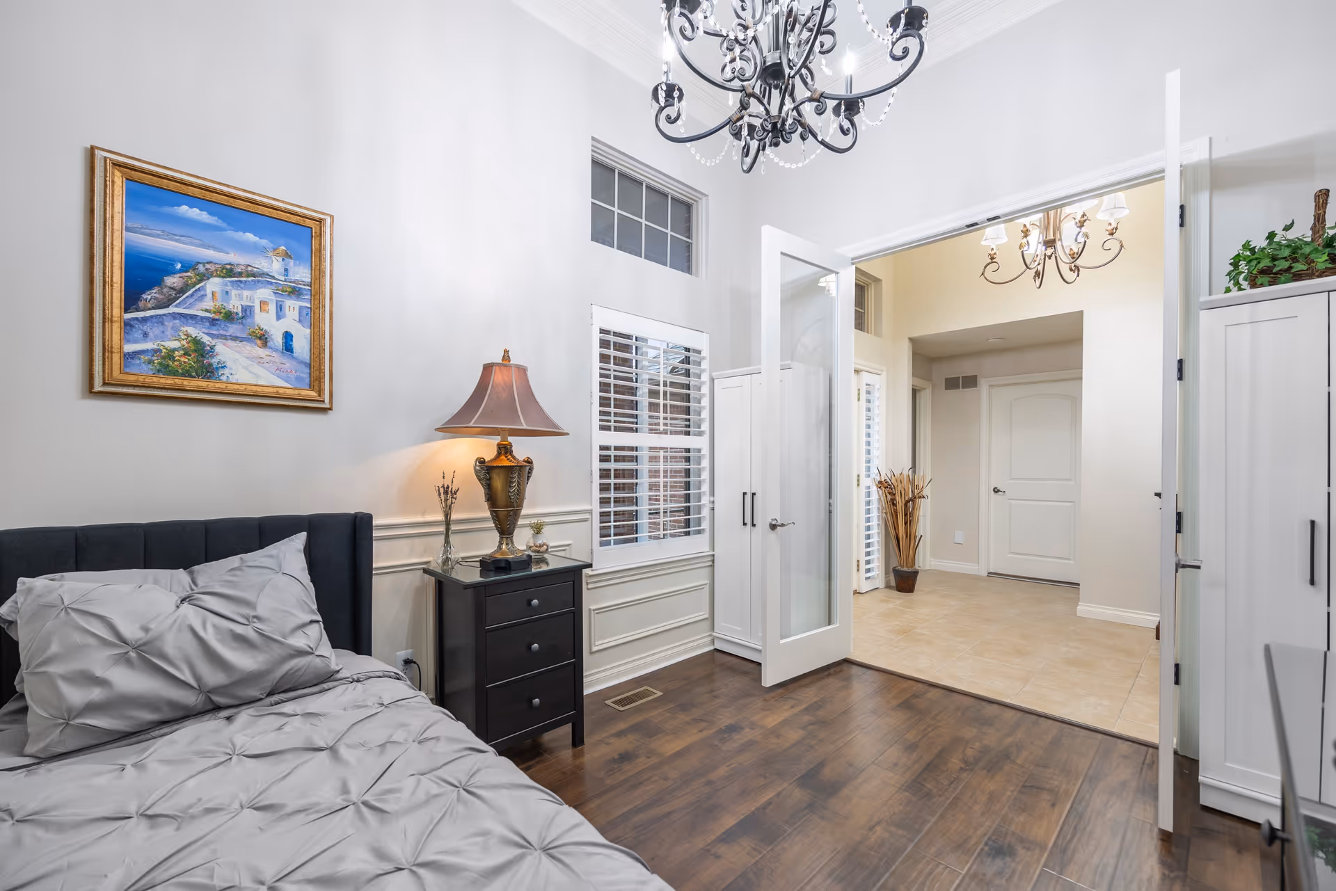 Bright, neatly decorated bedroom with a bed, nightstand and lamp, chandelier, and open double doors leading to a tiled hallway.