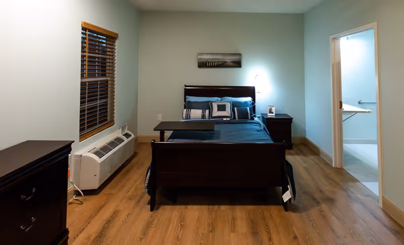 A neatly arranged bedroom with a wooden bed featuring blue bedding and multiple pillows. There is a bedside table with a lamp and a small framed photo. A window with wooden blinds is on the left wall above a white air conditioning unit. The floor is wooden, and an open door on the right leads to a bathroom with grab bars and a fold-down shower seat.