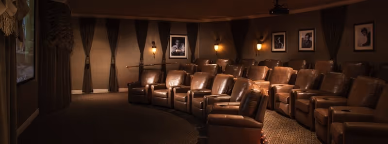 A cozy home theater room with multiple rows of brown leather recliner chairs facing a large screen. The walls are decorated with framed black and white portraits and have dark curtains. Warm wall sconces provide soft lighting.