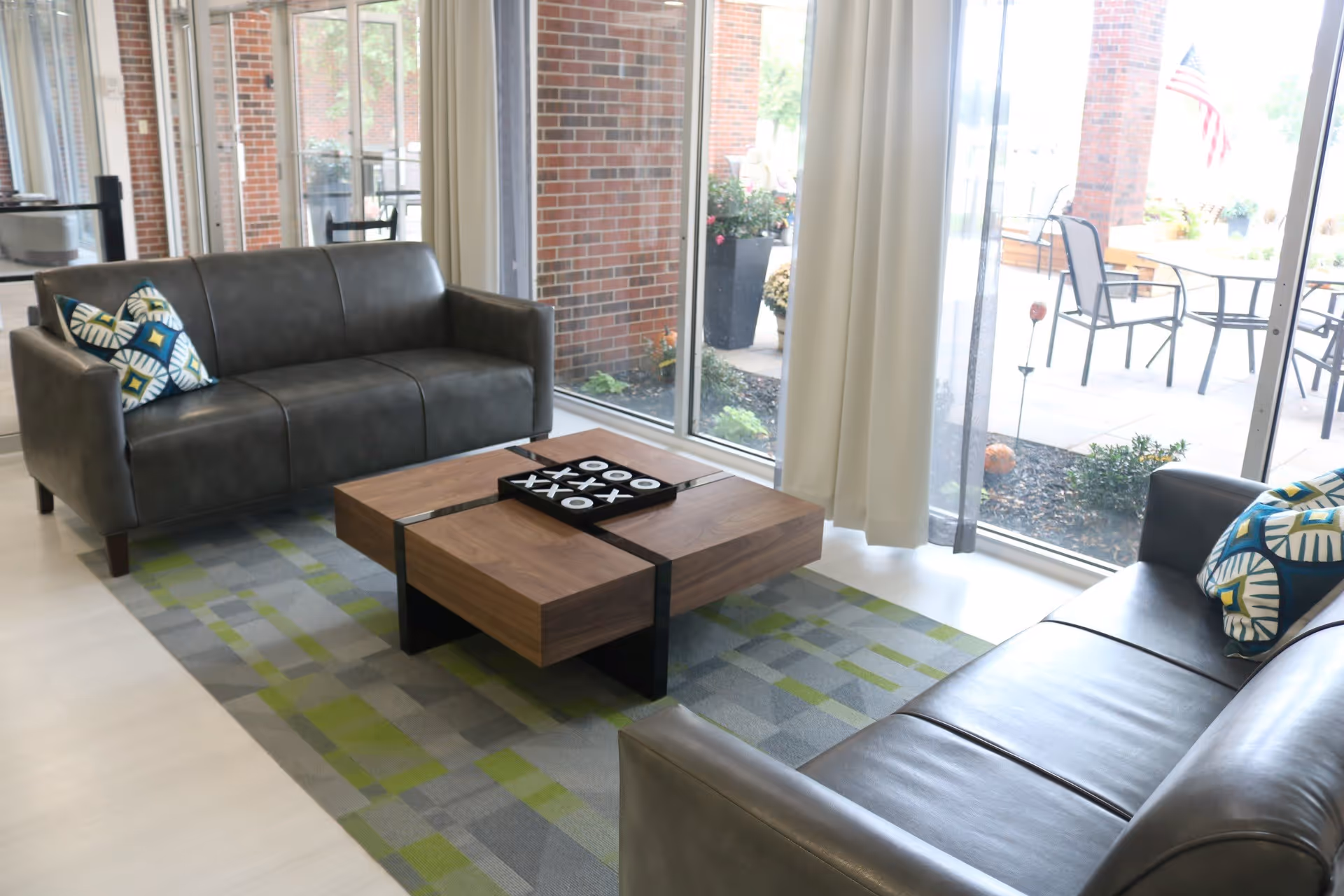 A bright living room area with two dark gray leather sofas facing each other, each with patterned throw pillows. A modern wooden coffee table with a tic-tac-toe game sits on a green and gray patterned rug. Large windows with white curtains provide a view of an outdoor patio with chairs and tables, and an American flag is visible outside.