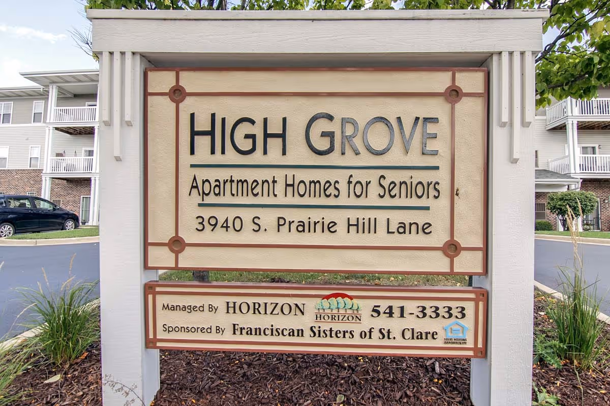 Sign for High Grove Apartment Homes for Seniors located at 3940 S. Prairie Hill Lane, managed by Horizon and sponsored by Franciscan Sisters of St. Clare, with a background showing part of the apartment building and parked cars.