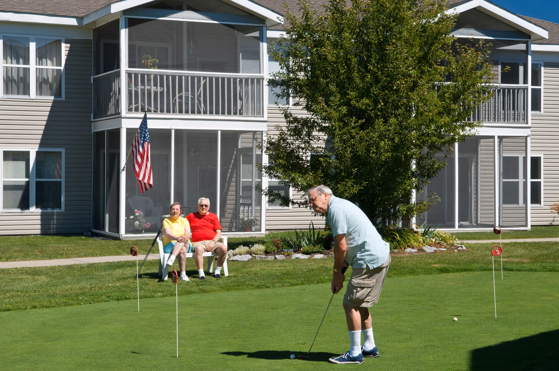 An elderly man is playing golf on a putting green outside a senior living facility. Two elderly people, a man and a woman, are sitting on white chairs nearby watching him. The building behind them has screened balconies and an American flag is displayed near the seated couple. There is a tree and some landscaping around the green.