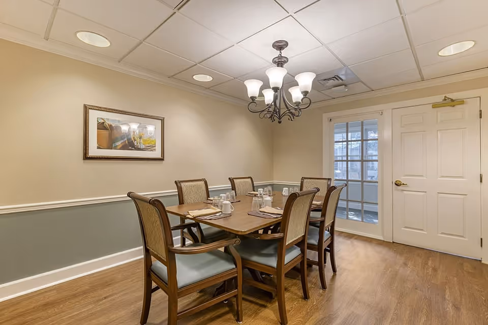A dining room with a wooden table set for six people, each place setting includes a napkin, cup, and glass. The room has beige walls with a green lower half, a framed picture on the wall, a chandelier with six lights hanging above the table, and a door with glass panels leading to another area.