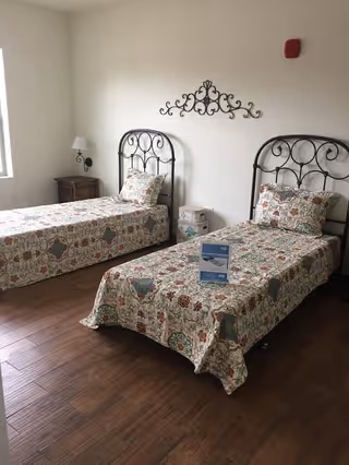 A room with two single beds featuring black metal headboards and floral patterned bedspreads. There is a wooden nightstand with a lamp between the beds, a decorative wall piece above the beds, and a window letting in natural light on the left side.