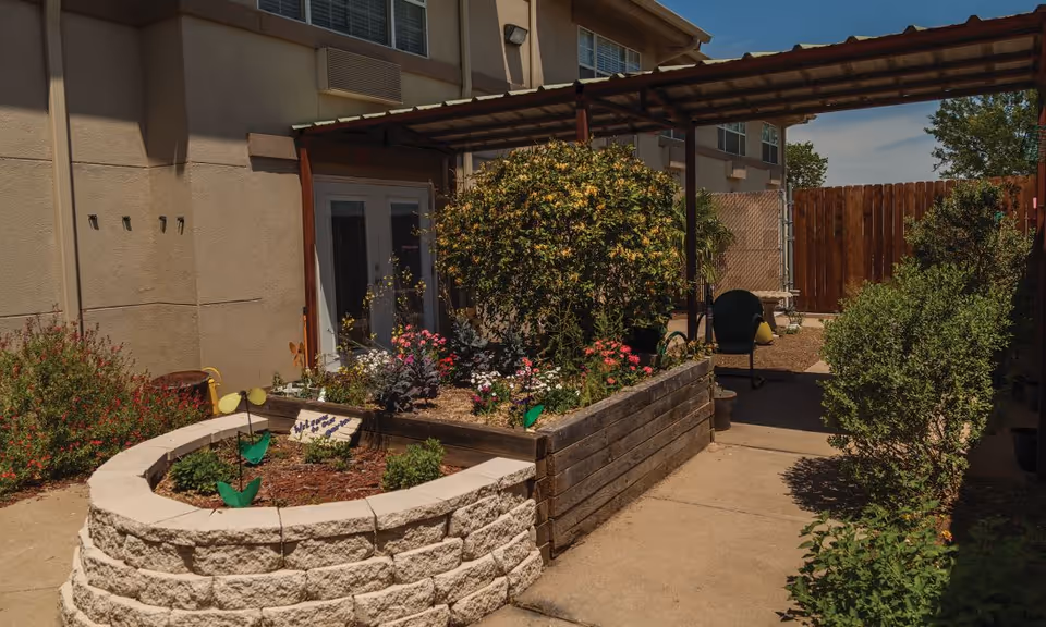 Outdoor garden area at Brayden Park Assisted Living & Memory Care featuring raised flower beds with various plants and flowers, a covered walkway, and a wooden fence in the background.