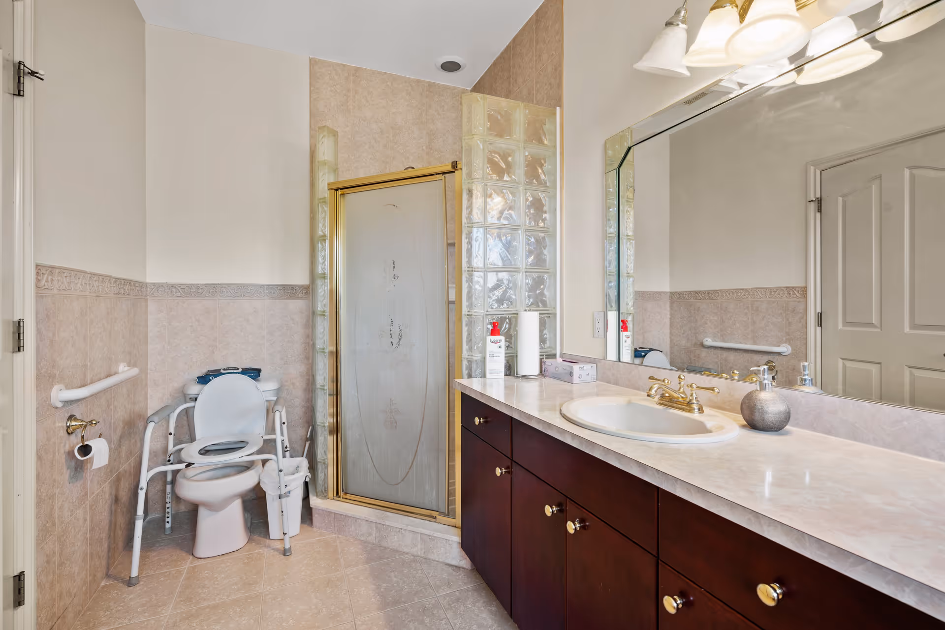 A bathroom with beige tiled walls and floor, featuring a toilet with a white safety frame around it, a glass-enclosed shower with a gold frame, a large mirror above a countertop with a sink, and various toiletries including lotion and a soap dispenser.